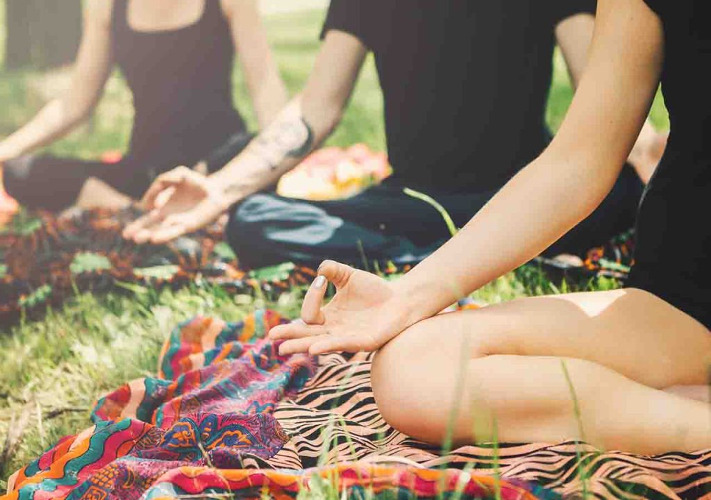 Three people, close up on their knees and hands, sit cross legged outdoors doing yoga.